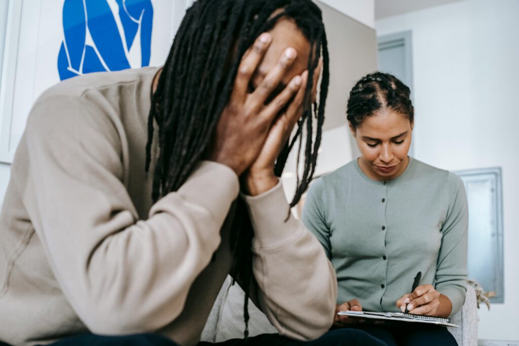 A black man with dreadlocks is holding his head in his hands while an African-American woman next to him takes notes