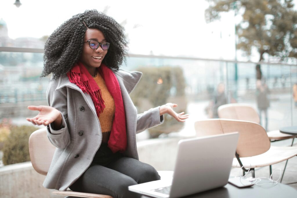 A black girl wearing a winter jacket on a terrace smiling while talking to someone on her laptop